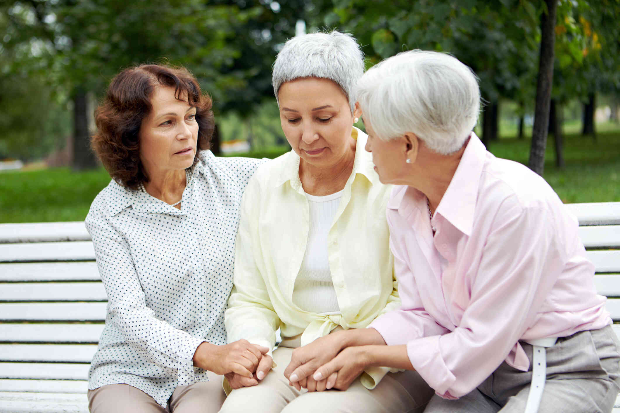 Three women sit together on a bench outdoors, holding hands and comforting the woman in the middle.