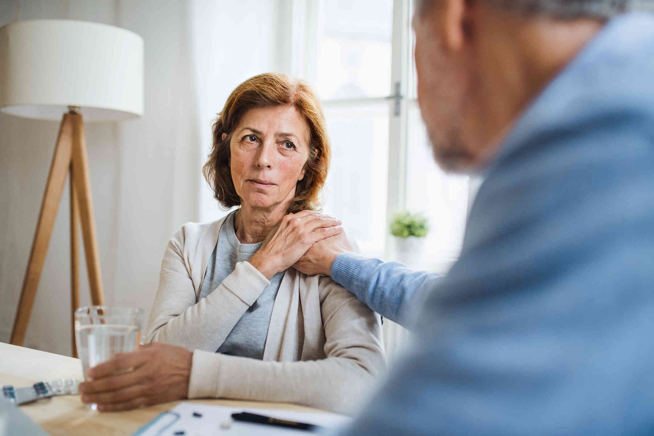 An elderly woman sits on a chair holding a glass of water in one hand and rests her other hand on her shoulder which an elderly man holds. She is looking sad. 