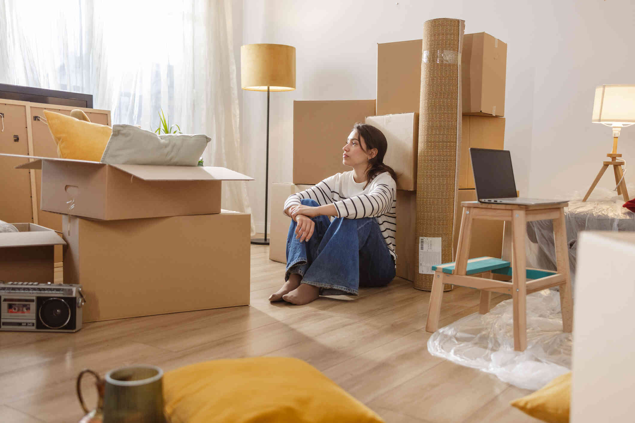 A woman sits on the floor surrounded by boxes. She appears lost in thought.