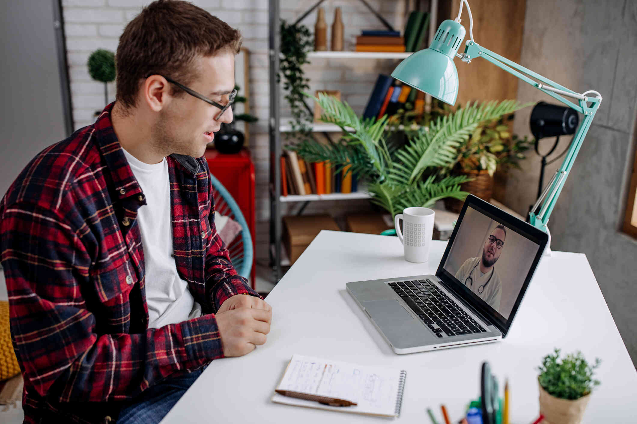 A man sits at a table talking to his therapist on his laptop