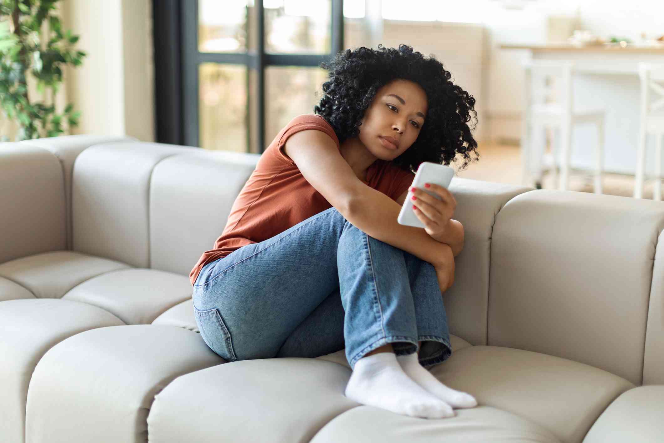 A woman seated on the couch, hugging her knee, is scrolling on her phone.