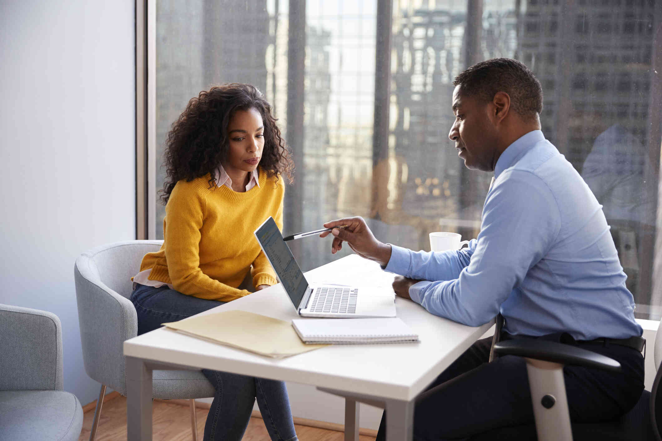 A man sits at a table with a laptop with a laptop in front of him speaking to a woman in a yellow shirt seated opposite him