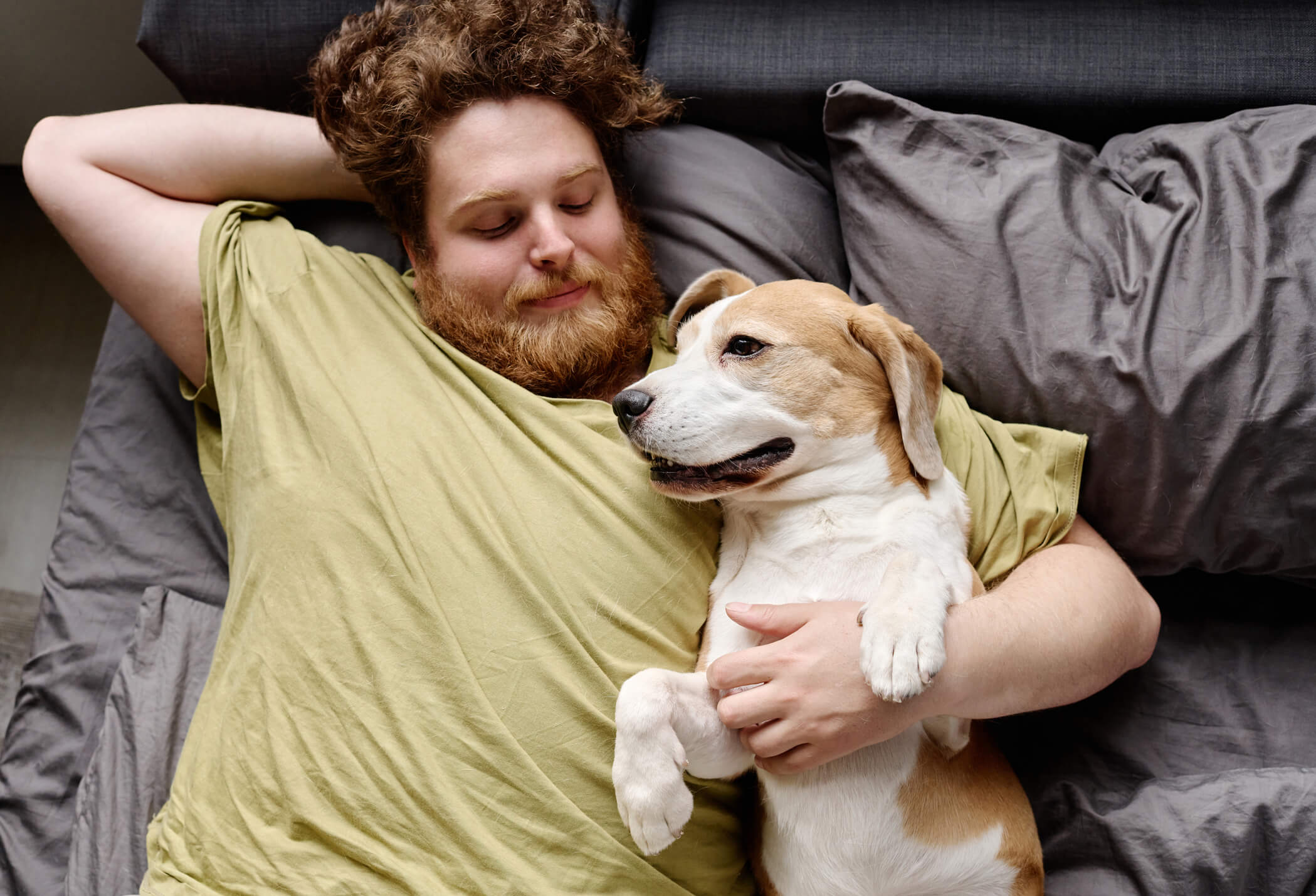 A man with curly brown hair, a beard, and a yellow-green t-shirt lies in bed and hugs his dog.